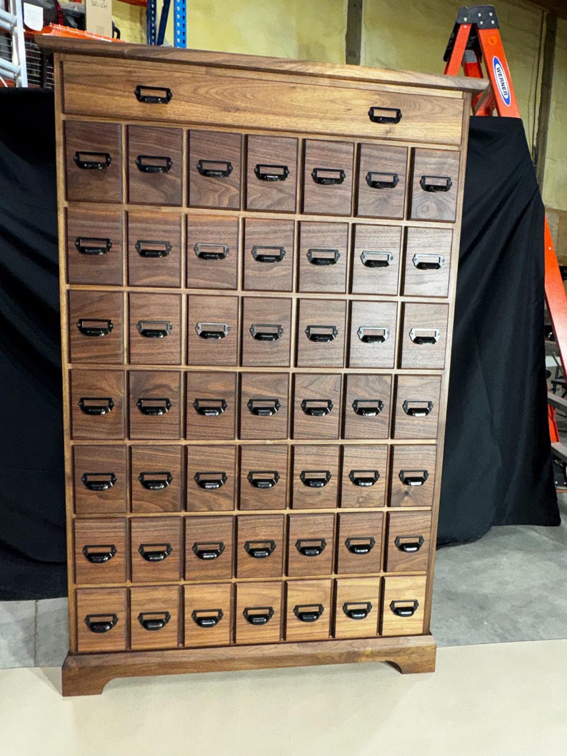 Puede incluir: Un mueble de boticario de madera con numerosos cajones peque&ntilde;os, cada uno con un tirador de metal. El mueble es de madera oscura y tiene forma rectangular. Los cajones est&aacute;n dispuestos en forma de rejilla y el mueble tiene un caj&oacute;n superior.