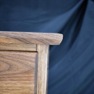 Puede incluir: Primer plano de un mueble de madera con acabado natural. El borde superior tiene una esquina redondeada. La veta de la madera es visible y el mueble parece estar bien elaborado. El fondo es azul oscuro.