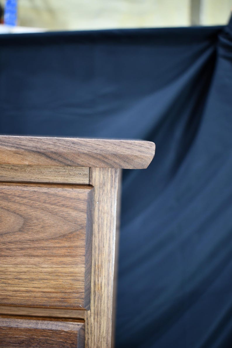 Puede incluir: Primer plano de un mueble de madera con acabado natural. El borde superior tiene una esquina redondeada. La veta de la madera es visible y el mueble parece estar bien elaborado. El fondo es azul oscuro.