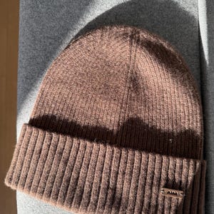 Könnte beinhalten: Eine braune Strickmütze mit geripptem Bund und kleinem Metallschild. Der Hut besteht aus einem weichen, strukturierten Material. Die Mütze hat eine neutrale Farbe, ist für verschiedene Outfits geeignet und soll den Kopf warm halten.