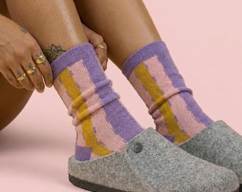 Könnte beinhalten: Graue Filzclogs mit einer lila, rosa und gelb gestreiften Socke. Die Socken sind hochgezogen und die Clogs haben eine runde Spitze und eine dunkle Sohle. Der Hintergrund ist einfarbig rosa.