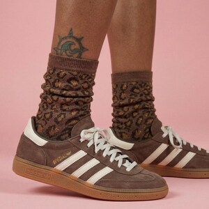 K&ouml;nnte beinhalten: Braune Adidas Spezial Sneaker mit wei&szlig;en Streifen und Schn&uuml;rsenkeln, kombiniert mit braunen Socken mit Leopardenmuster. Die Sneaker haben eine Gummisohle und das Wort "SPEZIAL" ist sichtbar. Der Hintergrund ist einfarbig rosa.