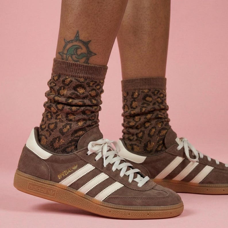 K&ouml;nnte beinhalten: Braune Adidas Spezial Sneaker mit wei&szlig;en Streifen und Schn&uuml;rsenkeln, kombiniert mit braunen Socken mit Leopardenmuster. Die Sneaker haben eine Gummisohle und das Wort "SPEZIAL" ist sichtbar. Der Hintergrund ist einfarbig rosa.