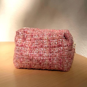 Puede incluir: Un neceser rectangular confeccionado en tejido de tweed rosa con textura. El material presenta un patrón tejido con hilos metálicos plateados entrelazados. Un pequeño tirador de cremallera de metal dorado es visible en el lado derecho.