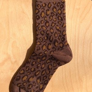 K&ouml;nnte beinhalten: Eine braune Socke mit einem Leopardenmuster in Braun- und Goldt&ouml;nen. Die Socke ist aus Strickmaterial gefertigt und wird auf einer Holzoberfl&auml;che gezeigt. Die Socke ist kn&ouml;chellang.