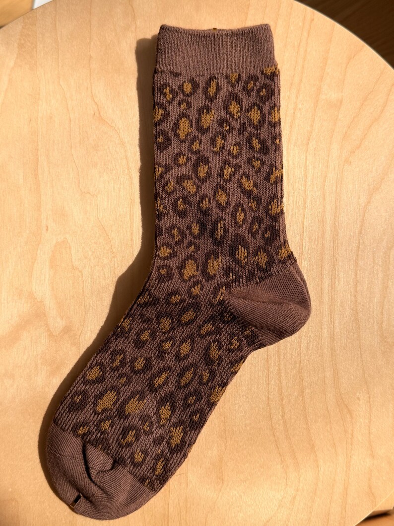 K&ouml;nnte beinhalten: Eine braune Socke mit einem Leopardenmuster in Braun- und Goldt&ouml;nen. Die Socke ist aus Strickmaterial gefertigt und wird auf einer Holzoberfl&auml;che gezeigt. Die Socke ist kn&ouml;chellang.