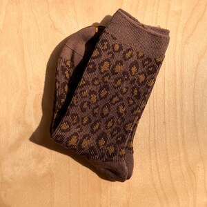 K&ouml;nnte beinhalten: Ein Paar braune Socken mit Leopardenmuster. Die Socken sind gefaltet und zeigen eine gestrickte Textur. Das Leopardenmuster kombiniert dunkleres Braun mit hellbraunen Flecken f&uuml;r einen stilvollen Look.