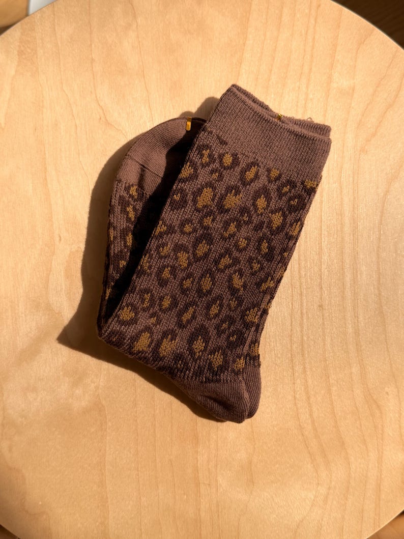 K&ouml;nnte beinhalten: Ein Paar braune Socken mit Leopardenmuster. Die Socken sind gefaltet und zeigen eine gestrickte Textur. Das Leopardenmuster kombiniert dunkleres Braun mit hellbraunen Flecken f&uuml;r einen stilvollen Look.