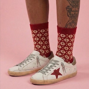 Peut inclure: Baskets blanches avec des lacets gris et une étoile rouge sur le côté, associées à des chaussettes rouges à motifs. Les chaussettes présentent un motif de losanges répétés dans une couleur plus claire. Les chaussures ont un aspect usé et vintage.