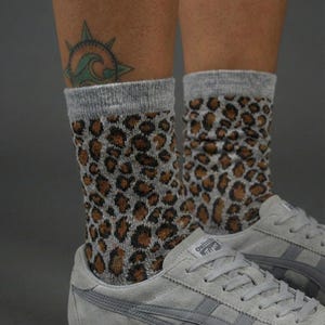 Op de afbeelding: Grijze sokken met een luipaardprint worden gedragen met grijze sneakers. De sokken hebben een grijze boord en een bruine en zwarte luipaardprint. De sneakers zijn grijs met witte veters en een witte zool.