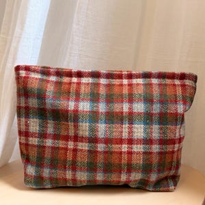 Puede incluir: Una bolsa rectangular de tela a cuadros en tonos rojos, naranjas, verdes y azules. La bolsa está hecha de un material texturizado y se encuentra sobre una superficie clara. El patrón es un diseño de cuadros clásico.