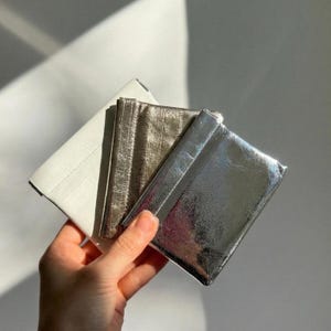 Op de afbeelding: Een hand die een stapel van drie kleine portemonnees vasthoudt in wit, metallic goud en zilver. De portemonnees hebben een glanzende, getextureerde afwerking. De witte portemonnee staat links, gevolgd door de gouden en zilveren portemonnees.