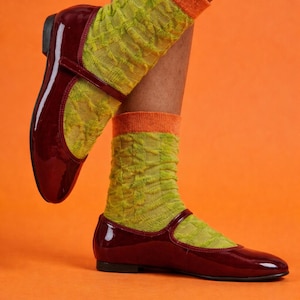 Könnte beinhalten: Lacklederne Mary Jane-Schuhe in Burgunderrot mit Riemen, kombiniert mit grün gemusterten Socken mit orangefarbenen Bündchen. Die Schuhe stehen vor einem leuchtend orangefarbenen Hintergrund.