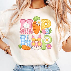May include: Cream-colored t-shirt with a colorful Easter-themed graphic. The design spells out "HOP HOP" with letters decorated as bunny feet, a carrot, and a bunny face wearing sunglasses. Easter eggs and flowers complete the design.