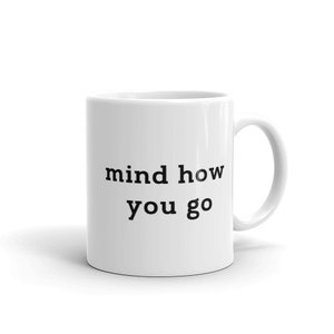 以下が含まれることがあります： 白いセラミック製のコーヒーカップで、黒い文字で「mind how you go」と書かれています
