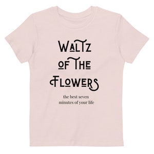 以下が含まれることがあります： 薄いピンク色のTシャツ。黒色の装飾的なフォントで「Waltz of the Flowers」と書かれています。その下に「the best seven minutes of your life」というフレーズがあります。