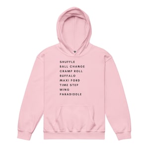 May include: A light pink hoodie with a front pocket and a hood. The text "SHUFFLE BALL CHANGE CRAMP ROLL BUFFALO MAXI FORD TIME STEP WING PARADIDDLE" is printed in black on the front. The hoodie is made of a soft material.