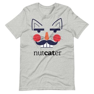 Op de afbeelding: Lichtgrijs t-shirt met een cartoon kattengezicht ontwerp. Het ontwerp bevat kattenoren, wenkbrauwen, rode wangen, een snor en het woord "nutcater" in zwarte tekst.