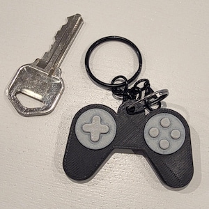 Puede incluir: Un llavero de mando de videojuego negro y gris con una llave plateada. El mando tiene una cruceta direccional y cuatro botones circulares. El llavero tiene un anillo y un cierre negros.