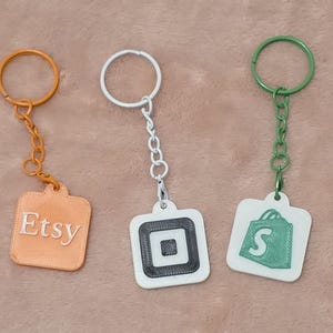Könnte beinhalten: Drei Schlüsselanhänger mit quadratischen Anhängern. Ein orangefarbener Schlüsselanhänger mit dem Wort "Etsy". Der zweite Schlüsselanhänger ist weiß mit einem schwarz-weißen quadratischen Design. Der dritte Schlüsselanhänger ist weiß mit einem grünen Einkaufstaschen-Design.