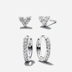 May include: A set of silver earrings. Two heart-shaped stud earrings with clear gemstones and two small hoop earrings with clear gemstones. The hoop earrings have the text "S925 ALE" on them.