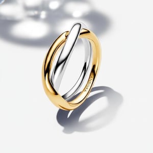 Könnte beinhalten: Ein zweifarbiges Ring mit einem goldfarbenen Band und einem silberfarbenen Band, das sich kreuzt. Der Ring liegt auf einer weißen Oberfläche und trägt die Gravur "PANDORA" an der Seite.