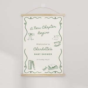 May include: A baby shower sign with the text "a New Chapter Begins" and "Welcome to Charlotte's BABY SHOWER". The sign is cream-colored with green decorative elements and is hanging from a wooden rod.