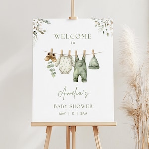May include: A baby shower sign on an easel with the words "Welcome to Amelia's Baby Shower" in green script. The sign features watercolor illustrations of baby clothes hanging on a clothesline, including shoes, a onesie, overalls, and a hat.