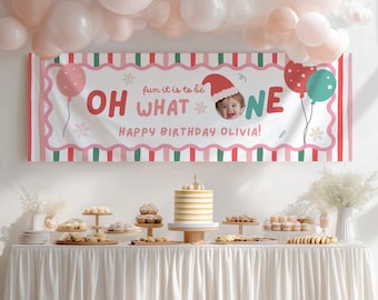 May include: A rectangular birthday banner featuring red and green stripes, a scalloped white border, and the text "OH WHAT FUN IT IS TO BE ONE" with "HAPPY BIRTHDAY OLIVIA!" below. It includes a photo of a baby in a Santa hat and balloons.