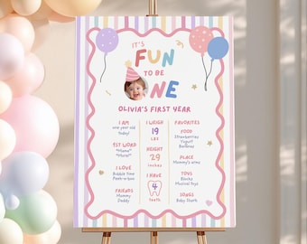 Op de afbeelding: Een rechthoekig verjaardagsbord met pastelkleurige verticale strepen en een geschulpte roze rand. De tekst luidt IT'S FUN TO BE ONE OLIVIA'S FIRST YEAR, inclusief babyfoto, gewicht van 19 lbs, lengte van 29 inches en 4 tanden.