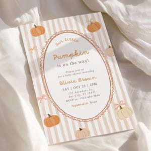 May include: A rectangular baby shower invitation card featuring a beige and white vertical striped background with orange pumpkin illustrations and small pink bows. The centre has an oval frame containing the text Our little Pumpkin is on the way!