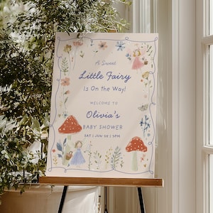 以下が含まれることがあります： 「A Sweet Little Fairy Is On the Way!」と「Welcome to Olivia's BABY SHOWER」の文字が入ったベビーシャワーのサイン。妖精、キノコ、花のイラストが描かれており、水色の縁取りと文字が特徴です。