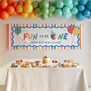 May include: A rectangular birthday banner with a white background, colorful striped borders, and the text "IT'S FUN TO BE ONE" with a photo of a baby wearing a party hat. The banner reads "HAPPY BIRTHDAY OLIVER" and is displayed above a table with a cake and treats.