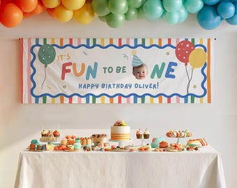 Op de afbeelding: Een rechthoekige verjaardagsbanner met een witte achtergrond, kleurrijke gestreepte randen en de tekst "IT'S FUN TO BE ONE" met een foto van een baby met een feesthoedje. De banner vermeldt "HAPPY BIRTHDAY OLIVER" en hangt boven een gedekte tafel.