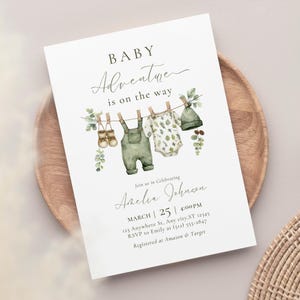 Könnte beinhalten: Babyparty-Einladung mit Aquarellillustrationen von Babykleidung, die an einer Wäscheleine hängt. Die Einladung enthält den Text "Baby Adventure is on the way" sowie Veranstaltungsdetails, einschließlich des Namens Amelia Johnson, Datum, Uhrzeit und RSVP-Informationen.