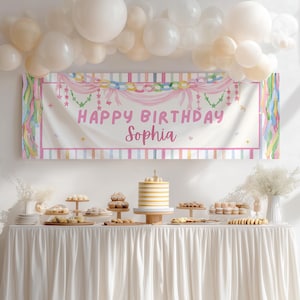 May include: A rectangular party banner featuring the text "HAPPY BIRTHDAY Sophia" in pink, decorated with pastel-colored streamers, hanging chains, and star accents. The banner is displayed above a table covered with a white cloth and various desserts.