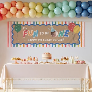 May include: A rectangular birthday banner with a tan center and colorful striped border, featuring the text IT'S FUN TO BE ONE HAPPY BIRTHDAY OLIVER, a photo of a baby in a party hat, and illustrations of balloons, displayed above a table with cake.