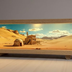 Pode incluir: Uma obra de arte retangular com moldura de madeira apresentando uma paisagem desértica com um edifício e dunas de areia. A obra de arte é iluminada, criando um brilho quente. A moldura é de cor clara e repousa sobre dois pequenos suportes de madeira.