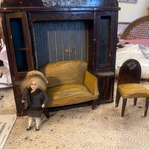 May include: Antique dollhouse furniture set featuring a dark wooden cabinet with a blue striped backdrop, a yellow sofa, and a small wooden chair. A doll in a gray outfit stands nearby.