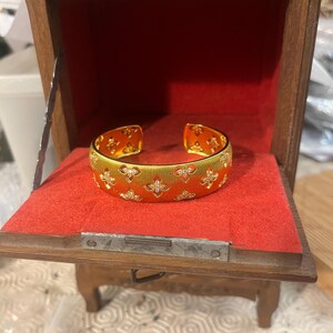 Peut inclure: Un bracelet manchette doré avec un motif floral et de petits pierres précieuses transparentes. Le bracelet est présenté dans un petit coffret à bijoux en bois avec un intérieur en velours rouge. La boîte est ouverte, exposant le bracelet.