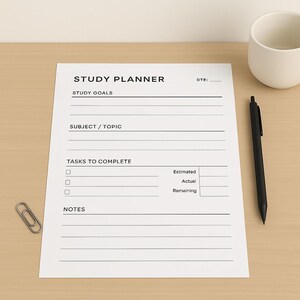Op de afbeelding: Een witte studieplanner met de titel "STUDY PLANNER" in zwarte tekst. De planner bevat secties voor studie-doelen, onderwerp/thema, taken en notities. Een zwarte pen en een zilveren paperclip liggen op een houten oppervlak.