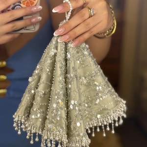 May include: A silver-coloured, beaded evening bag with a triangular shape. The bag is embellished with sequins and dangling pearl-like beads along the bottom edge. The bag is held by a person with long, manicured nails.