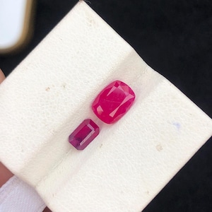 Könnte beinhalten: Zwei facettierte, rote Edelsteine unterschiedlicher Größe liegen auf einer weißen Oberfläche. Der größere Edelstein ist im Kissenschliff, der kleinere im Smaragdschliff. Die Edelsteine zeigen eine lebendige, gesättigte Farbe.