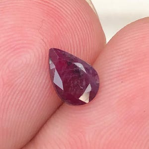 May include: A pear-shaped, faceted gemstone with a deep, saturated red color. The stone features multiple triangular and trapezoidal facets that reflect light, creating a brilliant appearance across its surface. The gem has a pointed top and a rounded base.