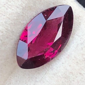 May include: A faceted, marquise-cut gemstone in a deep magenta colour. The polished surfaces of the gem reflect light, displaying its intricate cuts and internal brilliance. The gemstone is set against a white background, highlighting its colour and clarity.