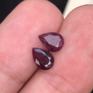 May include: Two pear-shaped, faceted rubies with a deep red colour and visible internal inclusions. The gemstones are cut with multiple triangular facets that reflect light, showcasing a dark, saturated crimson hue throughout the stones.