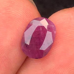 Peut inclure: Une pierre précieuse à facettes de forme ovale d'une couleur magenta-rose profond. La pierre présente une surface polie avec de multiples facettes triangulaires et trapézoïdales qui reflètent la lumière, créant un aspect translucide avec des inclusions.
