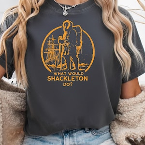 May include: Dark grey t-shirt with an orange circular graphic. The graphic depicts a figure with a backpack, a ship, and the text "WHAT WOULD SHACKLETON DO?". The design is a vintage-style illustration.