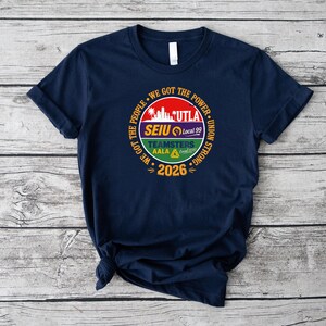 May include: Navy blue t-shirt with a circular graphic. The graphic includes the text "WE GOT THE PEOPLE" and "WE GOT THE POWER UNION STRONG". The graphic also includes the text "UTLA", "SEIU Local 99", "TEAMSTERS AALA Local 2010", and "2026".