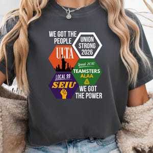 Op de afbeelding: Donkergrijs t-shirt met de tekst "WE GOT THE PEOPLE" en diverse logo's, waaronder "ULTA" met een skyline, "UNION STRONG 2026", "TEAMSTERS ALAA" en "SEIU" met een vuist.